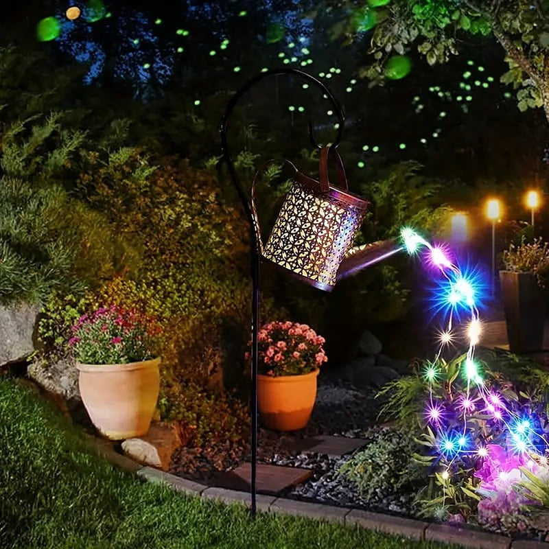 Multicolor watering can light on its stake beside a potted plant along a garden path at dusk
