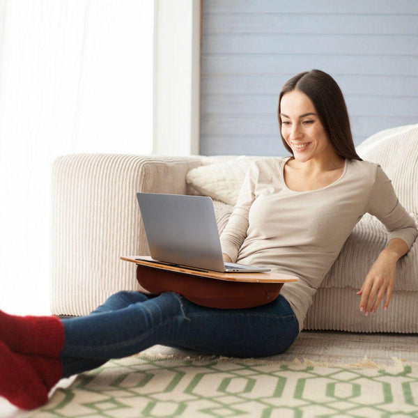 Smiling woman holding a laptop computer resting on a Bamboo Laptop Lap Desk with Pillow Cushion Stand Holder Table while sitting down on the floor with her back resting on a sofa couch